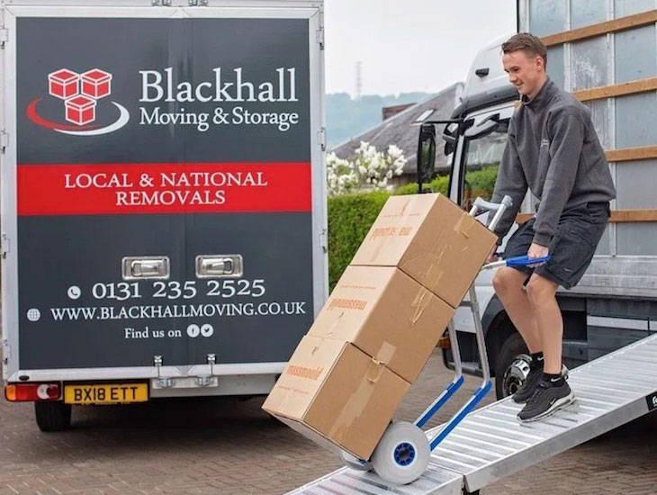 Young man pulling boxes on sack truck up a ramp onto removal van